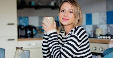 Jonge dame in trui zit aan tafel tijdens het ontbijt met een kop koffie in de keuken.